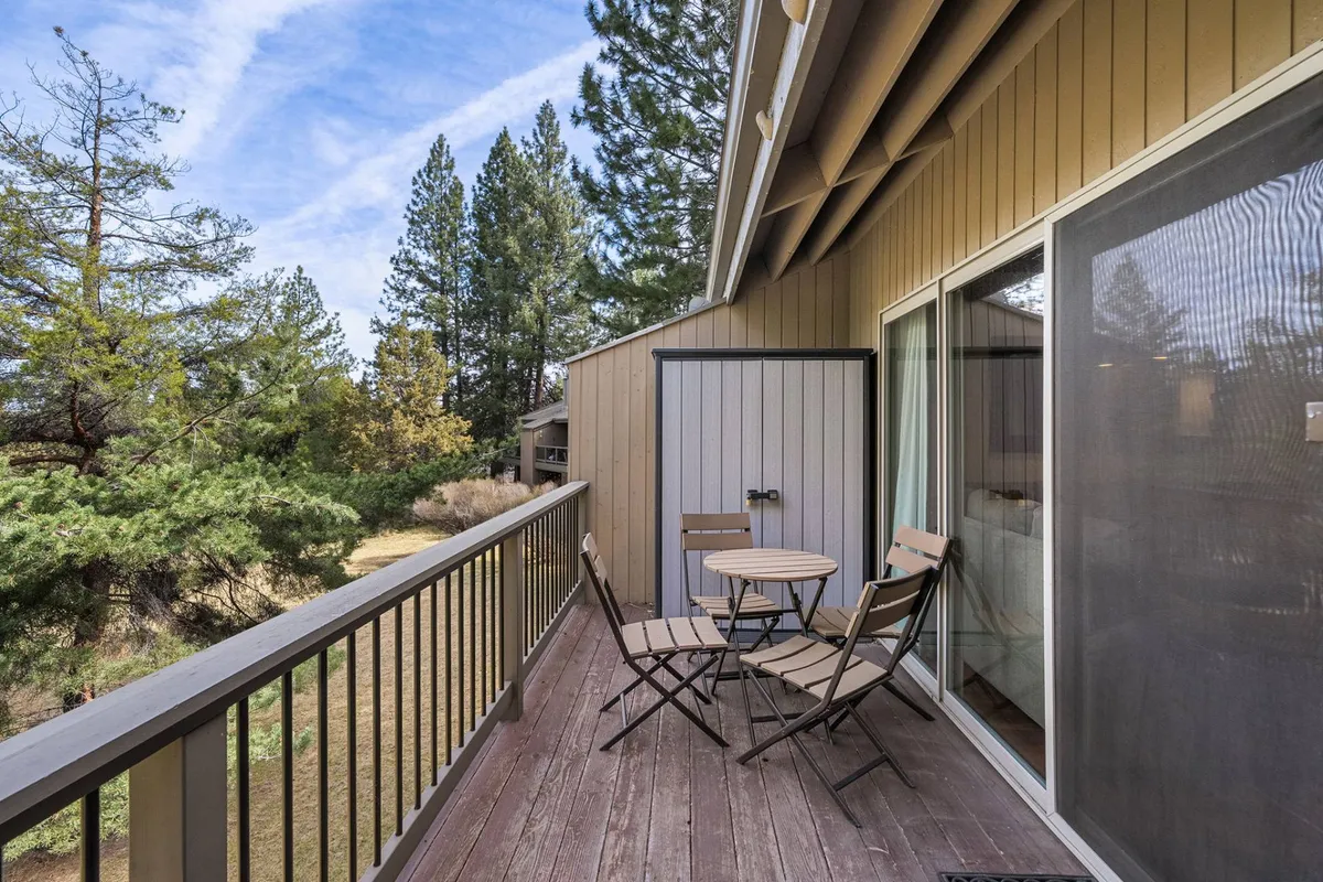 A cozy wooden balcony deck is furnished with a small round table and folding chairs, surrounded by lush green trees. The deck invites guests to enjoy quiet mornings, read a book, or share a conversation while taking in the natural beauty just outside the door.