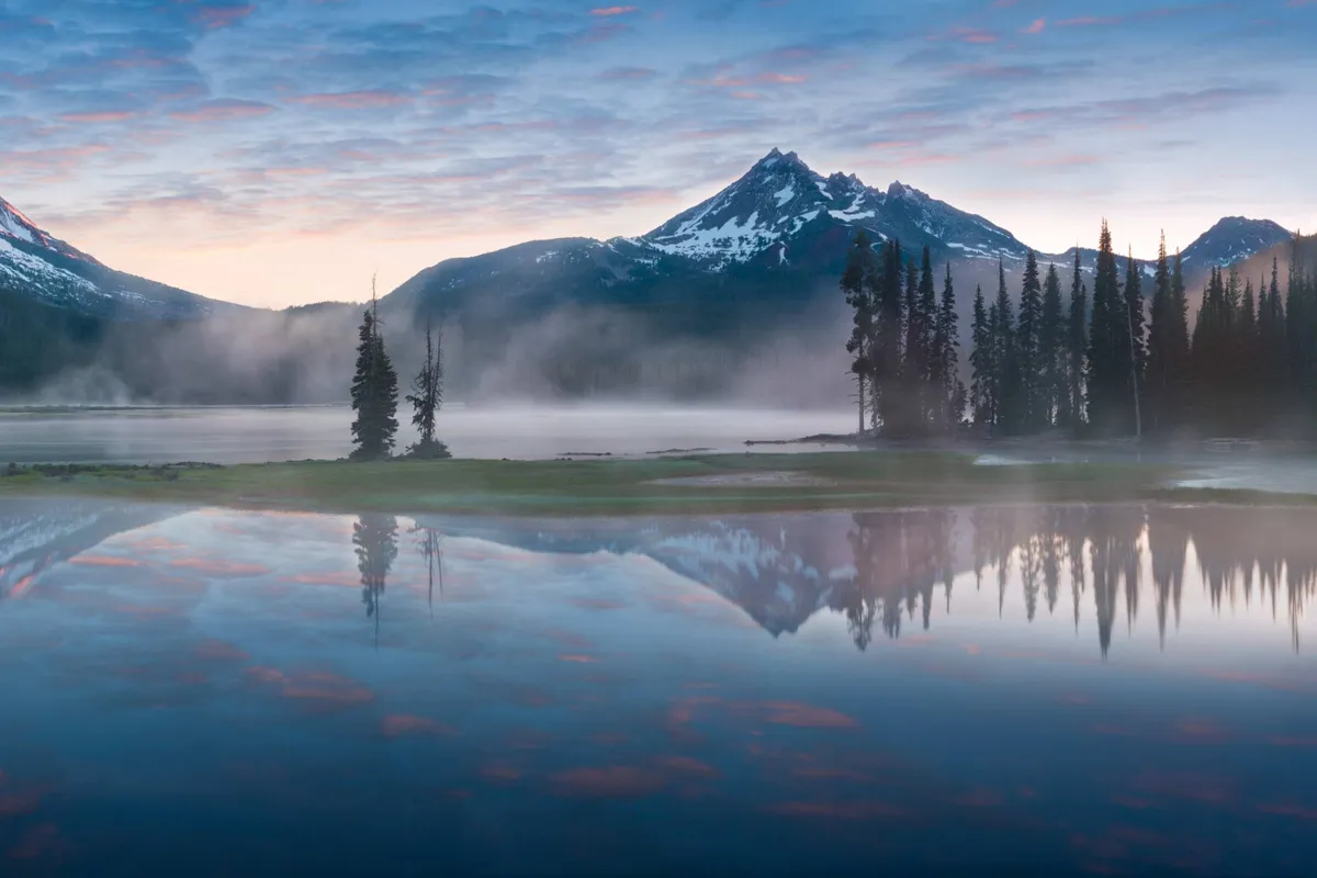 An expansive view encompassing misty mountains, reflective water, and a backdrop of trees. The scene feels tranquil and immersive, suggesting opportunities for hiking, kayaking, and photography in the area’s natural landscape.