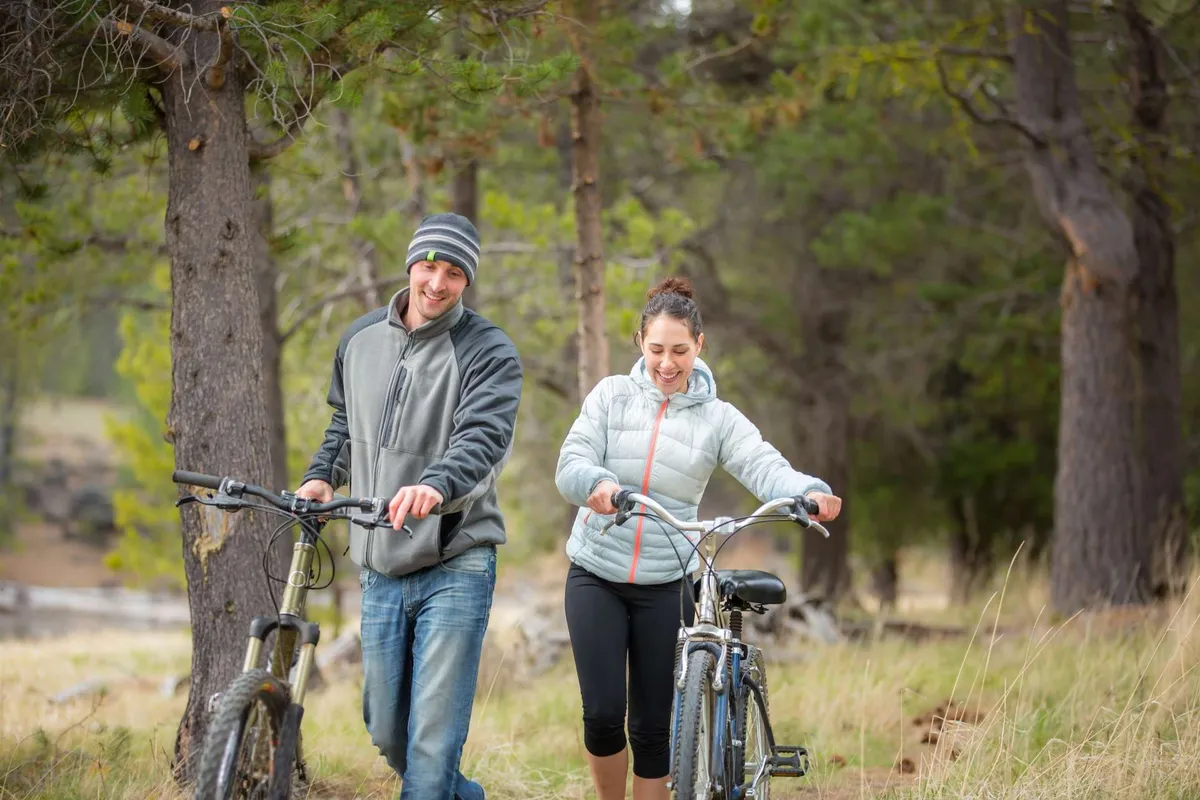 Two people walk side-by-side with bicycles along a trail surrounded by lush trees and a natural landscape, conveying active outdoor exploration and a connection to nature.
