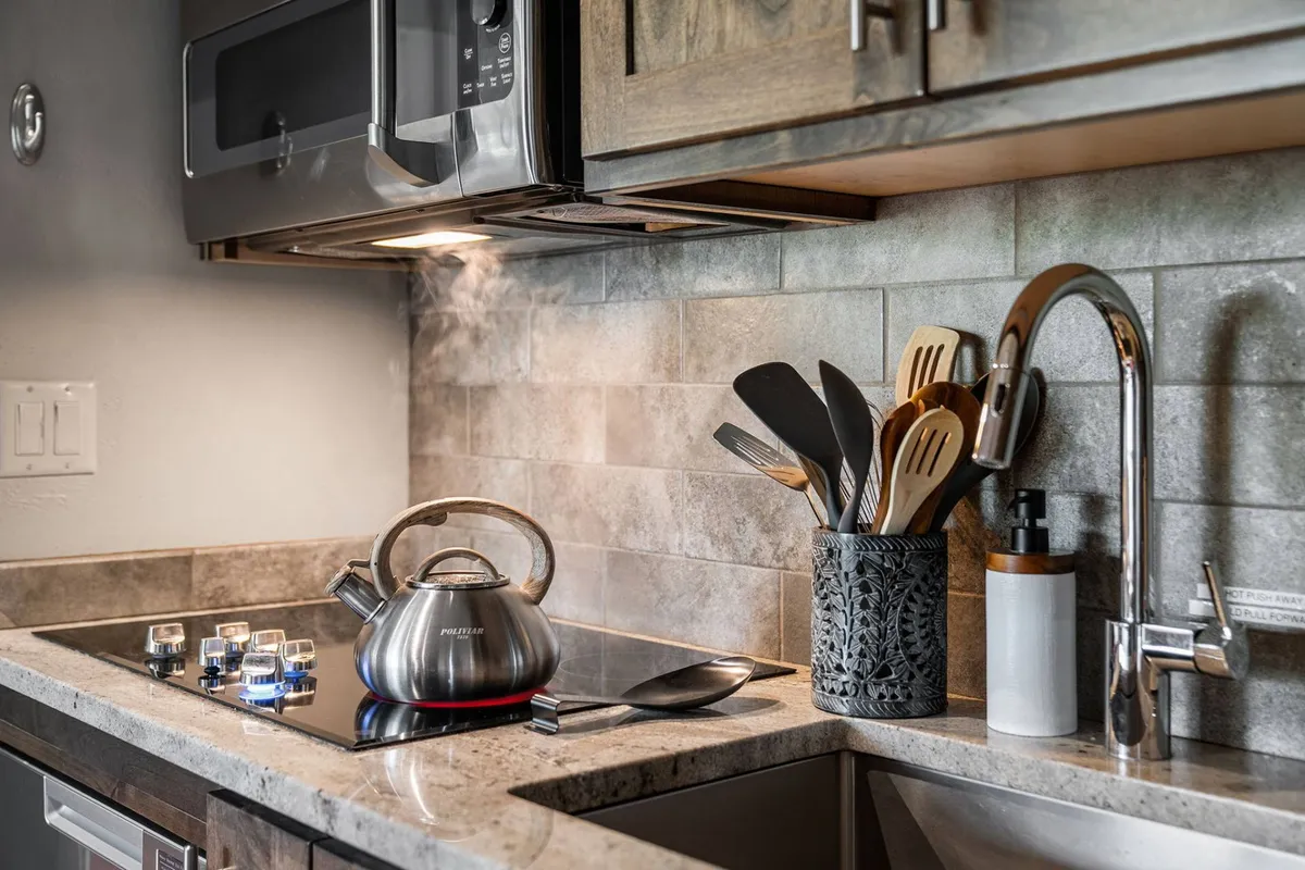 A close-up of a modern stove with blue flames, a stainless steel kettle, wooden utensils, and a sleek faucet set against a neat kitchen backdrop. The vignette evokes the anticipation and warmth of meal preparation, emphasizing the kitchen's functional details.