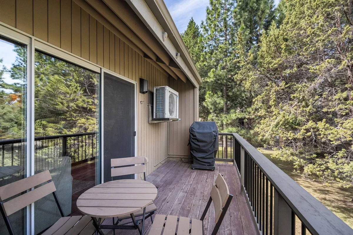 A peaceful exterior balcony features a small round table with wooden chairs, set against a backdrop of green foliage and an air conditioning unit. The scene is ideal for outdoor meals, quiet reflection, or sunset dinners.