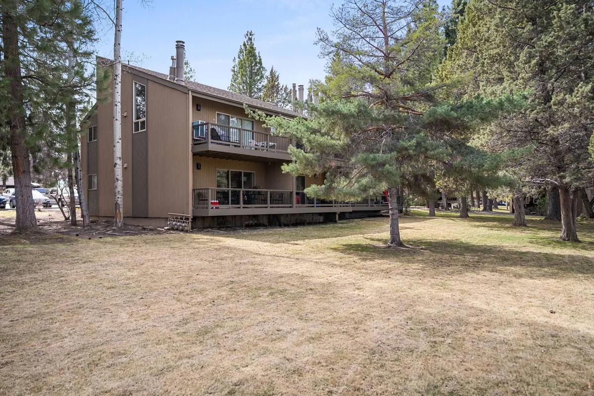 The two-story building features an inviting deck area surrounded by tall trees, showcasing the communal outdoor amenities of the resort. The deck is perfect for social gatherings, barbecues, or relaxing in nature.